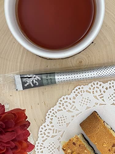 Assam Black Gold Tea stick on wood table beside white cup of brewed tea, doily, red flower, pastry slice