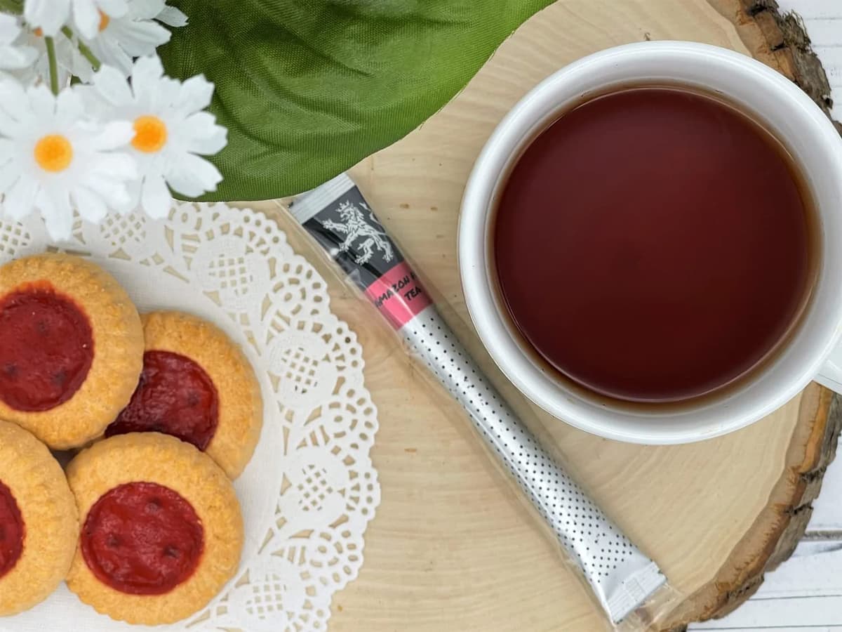 Amazon Fruit Tea stick on wood slice beside white cup of brewed tea, jam thumbprint cookies, white daisies