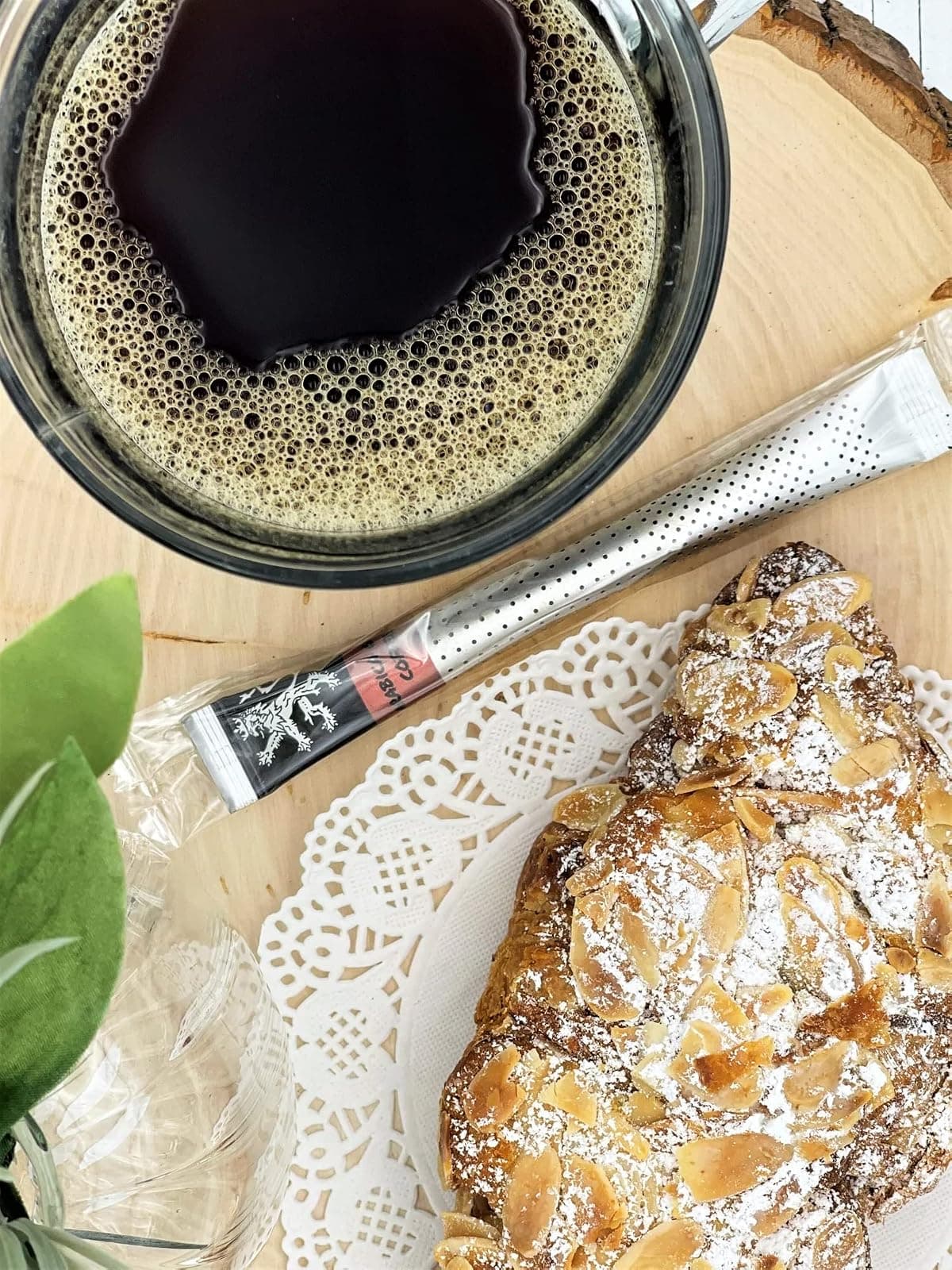 Arabica Gold Instant Coffee stick on wood slice beside glass mug of foamy brewed coffee, almond croissant, doily, foliage