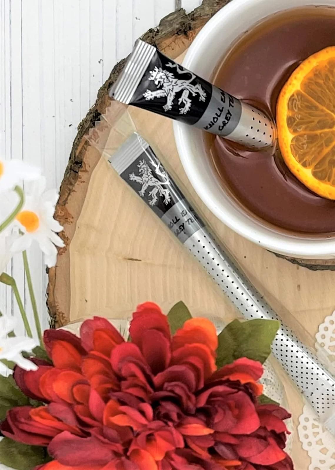 Knoll Earl Grey Tea stick two wrapped sticks on wood slice, white cup of brewed tea with orange slice, daisies, red and orange flowers