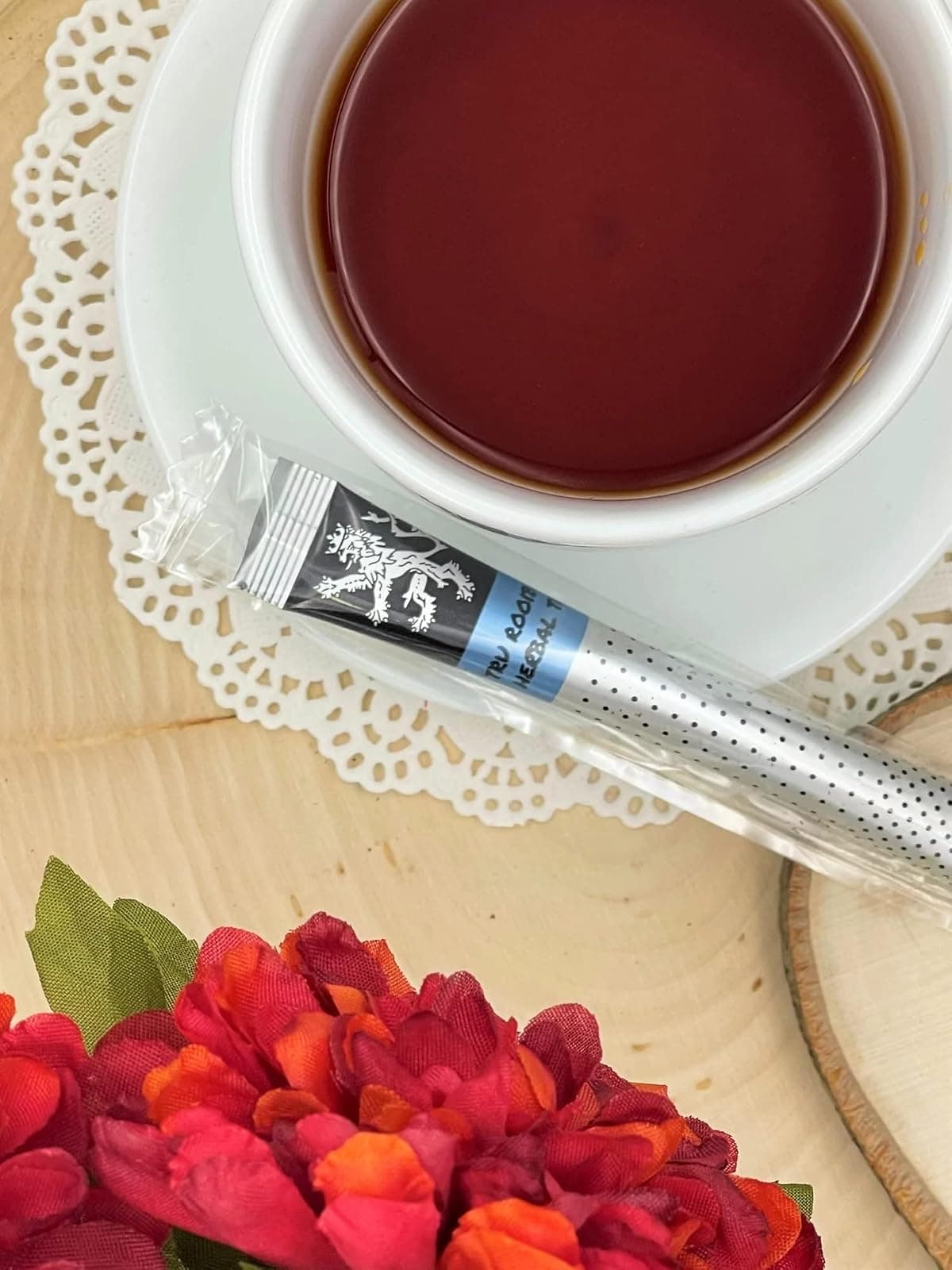 Tru Rooibos Tea stick on wood board and doily beside white cup+saucer of brewed tea, red and orange flowers